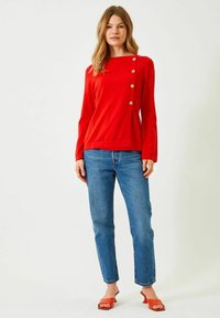 Punainen pitkähihaistoppi, jossa on neliömäinen pääntie ja koristeelliset kultaiset napit toisella puolella, yhdistettynä suoraan jalkaan meneviin sinisiin farkkuihin ja punaisiin sandaaleihin.