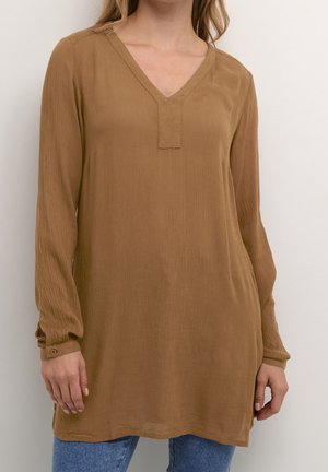 Kvinde iført en løs brun langærmet tunika med V-hals og blå jeans, stående mod en ensfarvet hvid baggrund.