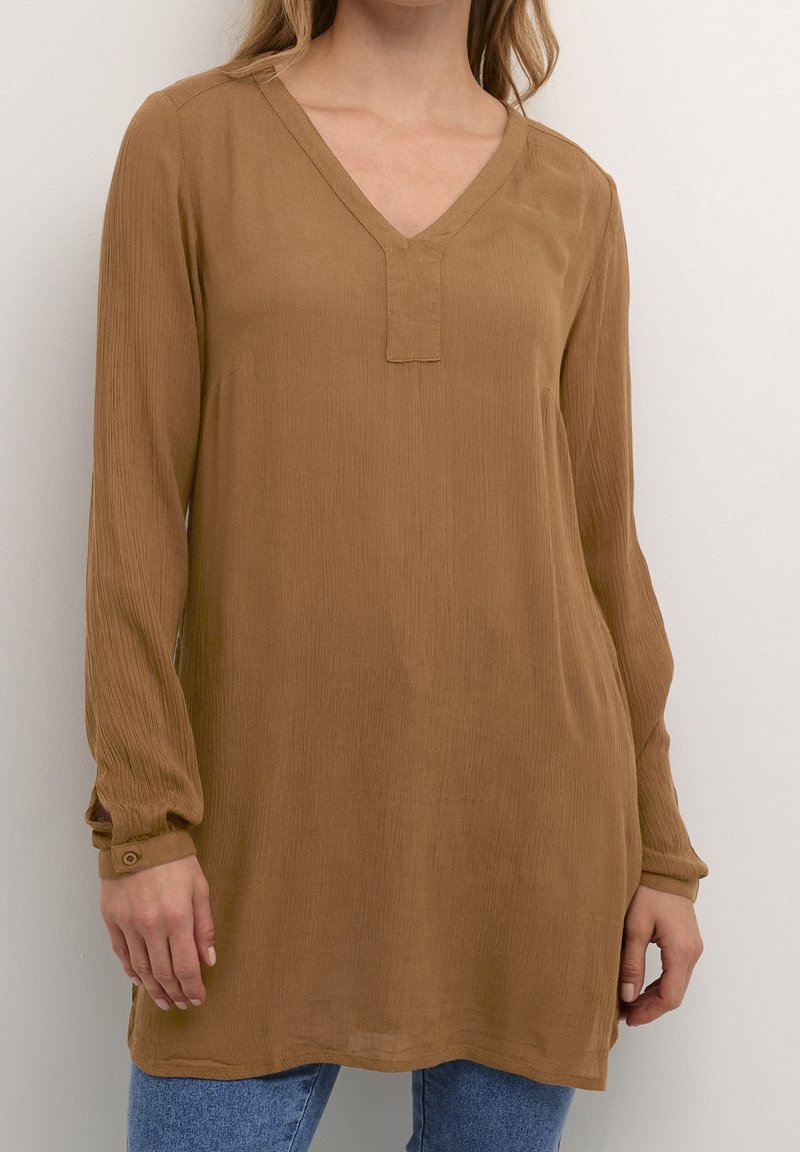 Femme portant une tunique ample marron à manches longues avec un col en V et un jean bleu, debout devant un fond blanc uni.