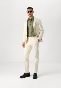 Beige dress med en strukturert design, kombinert med en olivengrønn skjorte og svarte sko. Figuren har på seg mørke solbriller.