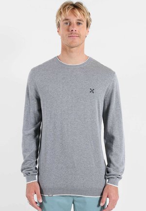 Homme blond portant un pull uni gris clair à manches longues avec un petit logo noir sur la poitrine et un pantalon bleu clair.