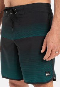 Sorte badshorts med teal horisontale striper, elastisk midjebånd med snor, og en liten logo-patch på nederste kant.