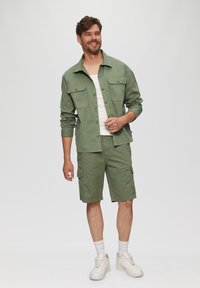 Groene katoenen shirt met twee borstzakken; bijpassende groene cargoshorts; witte tanktop; witte sneakers; relaxte pasvorm.