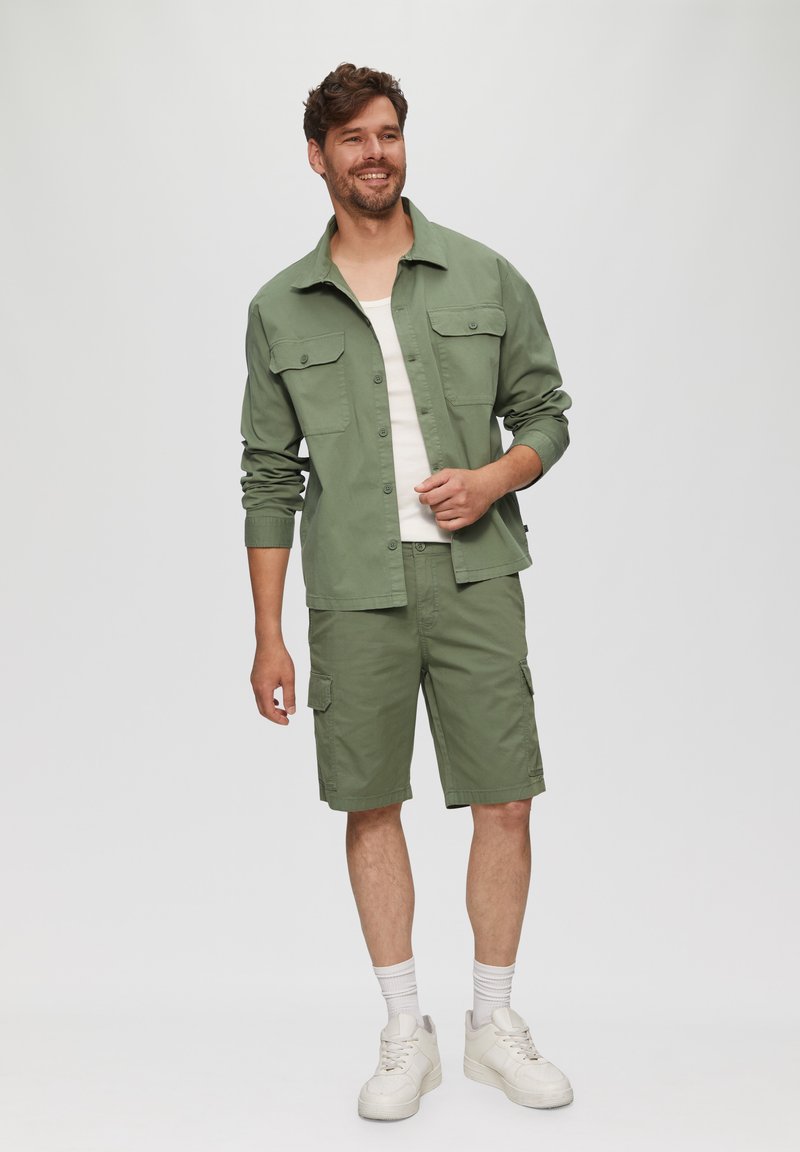 Groene katoenen shirt met twee borstzakken; bijpassende groene cargoshorts; witte tanktop; witte sneakers; relaxte pasvorm.