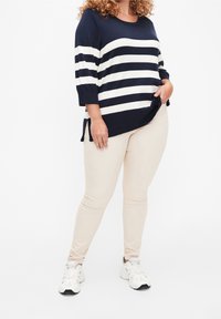 Marineblauwe en witte gestreepte gebreide trui met driekwart mouwen, gecombineerd met beige slim-fit broek en witte sneakers. Zijsplits aan de trui.