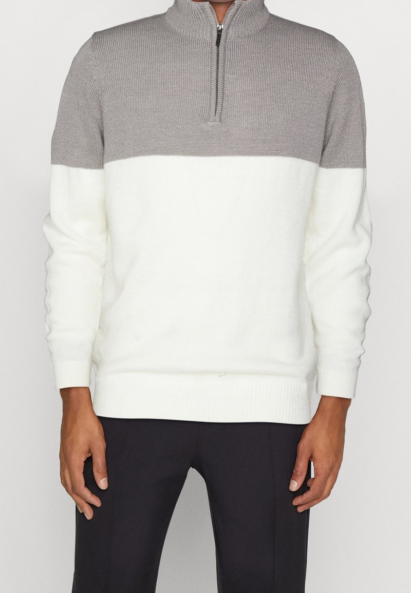 Homme portant un pull bicolore à fermeture éclair quart, avec le haut gris et le bas blanc, associé à un pantalon noir, debout devant un fond uni.