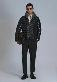 Padded schwarzes Jacket mit glanzender Oberfläche, mit einem mit Fell gefütterten Kragen, kombiniert mit dunklen Anzughosen und schwarzen Schnürschuhen. Schwarze Tasche in der Hand.