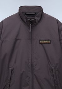Grå zip-up jakke med høj krave, sidelommer og et rektangulært mærke med sort og guld tekst. Glat stofstruktur.