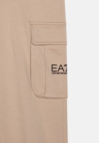 Shorts cargo beiges en coton doux, avec une poche à rabat sur le côté. Logo noir "EA7 Emporio Armani" imprimé sur la poche.