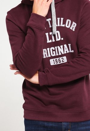 Person trägt einen kastanienbraunen Hoodie mit grauen Kordeln und weißem Text "TOM TAILOR LTD. ORIGINAL 1962", Arme verschränkt und Hand nahe am Gesicht.