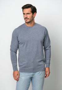 Maglione grigio in un morbido tessuto a maglia con maniche lunghe, scollatura rotonda e polsini e orlo a coste. Indossato con jeans azzurri.