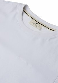 Bela bombažna majica s krožnim izrezom, ki ima subtilno izbočeno logotip na sprednji strani in teksturirano kremno oznako znotraj.