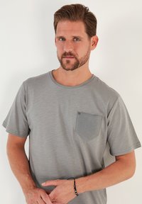 Siva kratka majica s kratkimi rokavi z teksturirano žepno podrobnostjo na prsih, okroglo ovratnikom in mehkega materiala, ki ima subtilen heathered vzorec.