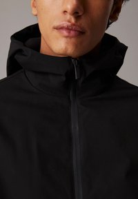 Giacca nera con cappuccio dal texture morbido, caratterizzata da una zip a lunghezza intera e un design minimale. Il tessuto appare flessibile e leggero.