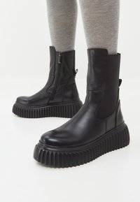 Botas de tobillo negras con una suela gruesa acanalada, cierre lateral con cremallera y paneles elásticos, con parte superior de cuero suave y punta redonda.