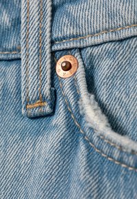 Lichtblauwe denimstof met een koperen knoop waarop "DAY" is gegraveerd, naast bruine stiksels en een textuurafwerking.