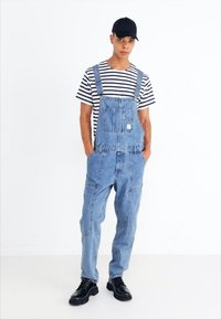 Salopette en denim bleu clair avec des bretelles ajustables, grande poche avant et poches latérales ; associée à un t-shirt à rayures noir et blanc et des chaussures noires.