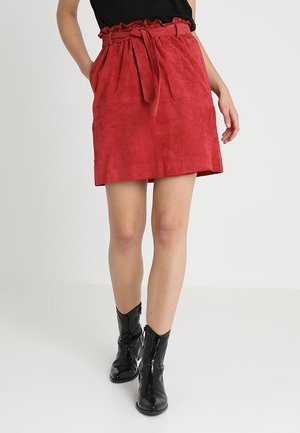 Kobieta nosząca czerwoną, zamszową spódnicę z wysokim stanem, z przednimi kieszeniami i paskiem do wiązania, połączoną z czarnymi skórzanymi butami sięgającymi do połowy łydki.