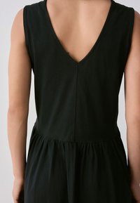 Sort ærmeløs kjole med V-formet ryg, flydende form, indsnævret talje og blød tekstur, der fremhæver et minimalistisk design.