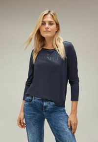 Dunkelblaues Langarm-T-Shirt mit rundem Halsauschnitt und dem Wort "CONNECT" auffällig präsentiert. Getragen mit hellblauen, modisch zerrissenen Jeans.