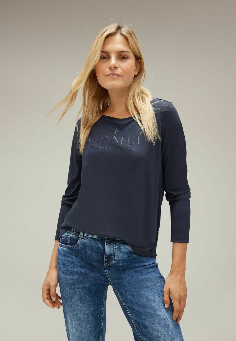 Dunkelblaues Langarm-T-Shirt mit rundem Halsauschnitt und dem Wort "CONNECT" auffällig präsentiert. Getragen mit hellblauen, modisch zerrissenen Jeans.