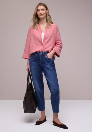 Jeune femme portant une chemise rouge à carreaux boutonnée, un jean bleu, des ballerines marron, tenant un grand sac fourre-tout marron foncé.