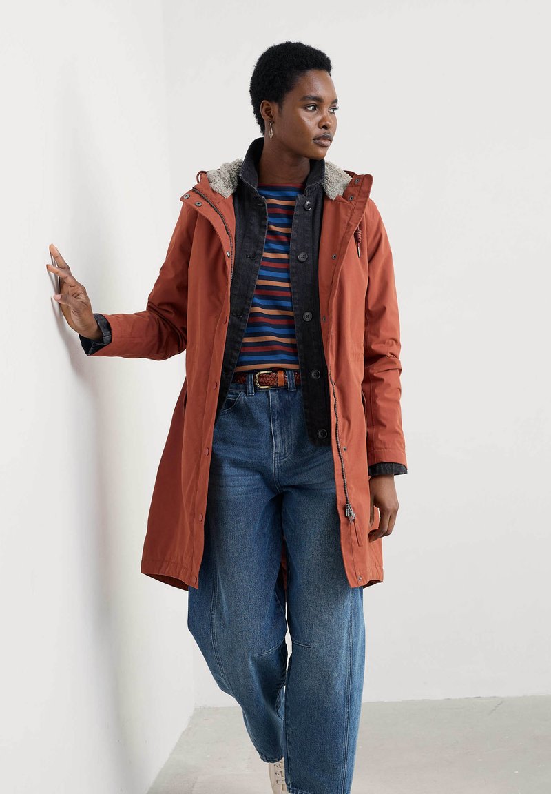 Orange utomhusjacka med fleece-fodrad huva, svart krage och framknappar, bärs över en randig skjorta med blå jeans.