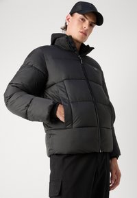 Svart pufferjakke med høy krage og frontglidelås. Har horisontal quiltedesign, to sidelommer og teksturert stoff.