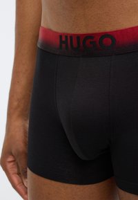 Boxer noir en matière douce avec une ceinture élastique dégradée rouge et noire portant le nom de la marque "HUGO" en lettres noires.