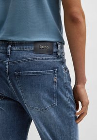 Blue denim jeans with a leather BOSS label on the back, featuring a slim fit, subtle fading, and standard pockets. Textured fabric surface.