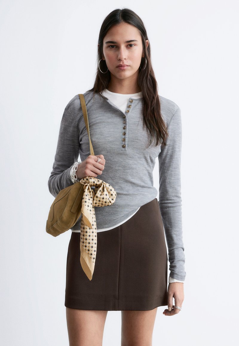 Kobieta o długich ciemnych włosach, ubrana w szary zapięty na guziki top, brązową spódnicę, kolczyki-koła, trzyma brązową torebkę z wzorzystą chustką.