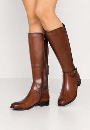 Gambe con stivali di pelle marrone al ginocchio, con cerniera laterale, cinturino decorativo e tacchi bassi a blocco su sfondo chiaro.