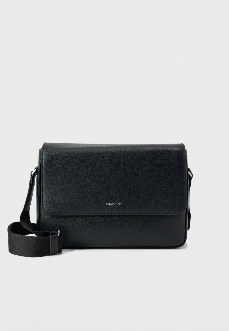 Μαύρη δερμάτινη τσάντα ταχυδρόμου με υφή φινίρισμα, ρυθμιζόμενο λουρί και μπροστινό καπάκι με διακριτικό λογότυπο. Συμπαγής και ορθογώνιο σχήμα.