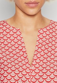 Red top featuring a wave pattern in white, with a v-neck design and textured fabric. Detail includes a button at the neckline.