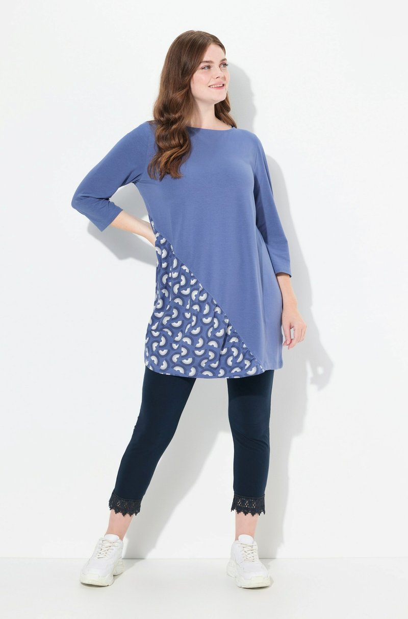 Túnica azul con mangas tres cuartos, panel lateral con estampado en blanco y leggins navy con detalle de encaje en el dobladillo. Zapatillas blancas.