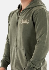 Haut à capuche zippé couleur olive fabriqué en tissu doux, avec un logo sur le devant, des poches kangourou et des poignets côtelés pour un maintien confortable.