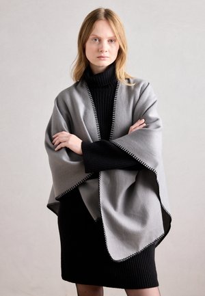 Junge Frau mit blondem Haar, die ein schwarzes Rollkragenkleid und einen hellgrauen Schal mit schwarzer Einfassung trägt, steht mit verschränkten Armen.