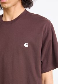 Barna pólót viselő személy, a bal felső mellen egy apró, fehér Carhartt logóval, fehér háttér előtt.