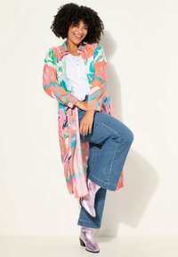 Kimono long et léger avec un motif abstrait multicolore, porté sur une chemise blanche et associé à un jean en denim évasé et des bottes roses brillantes.