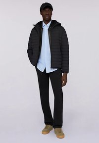 Svart dunjacka med horisontella sömmar, huva och dragkedja, bärs över en ljusblå randig skjorta och svarta byxor. Beige sneakers.