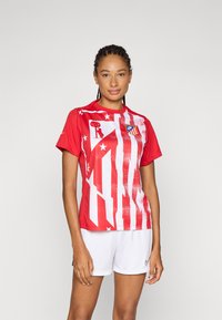 Rød atletisk trøye med hvite vertikale striper og et grafisk motiv, med klubbens logo på venstre bryst. Glatt stoff, rund halsdesign.
