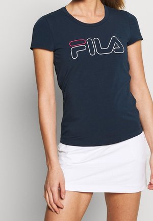 Frau trägt ein dunkelblaues FILA-T-Shirt und einen weißen Rock und posiert mit einer Hand entspannt an der Seite vor einem einfarbigen Hintergrund.