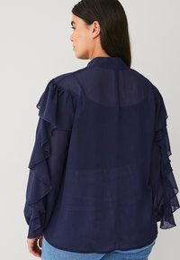 Kvinde med langt mørkt hår iført en gennemsigtig mørkeblå bluse med flæseærmer, stående sidelæns mod en ensfarvet lys baggrund.