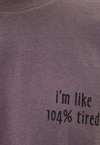 Lila bomullströja med svart text som säger "jag är typ 104 % trött", med rund halsringning och mjukt, texturerat tyg.