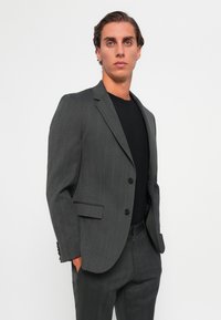 Grijs colbert met een subtiel visgraatpatroon, slim fit, met gelapte revers, twee knopen en één borstzak; gecombineerd met een zwart shirt.