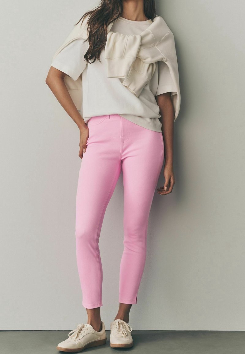 Femme portant une chemise beige à manches courtes avec un pull assorti drapé sur les épaules, un pantalon slim rose et des chaussures blanches décontractées.