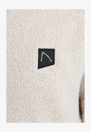 Tejido de forro polar beige difuso con una superficie texturizada. Presenta un parche negro con un diseño geométrico blanco, situado en la parte delantera.