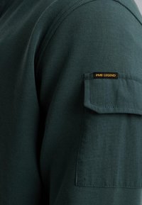 Dark green cotton long-sleeve shirt featuring a utility pocket on the left arm with a small black and yellow label reading "PME Legend."