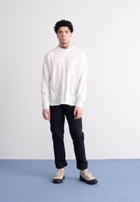 Wit overhemd met lange mouwen en ronde hals, gecombineerd met zwarte broek en beige schoenen. Eenvoudig ontwerp met subtiele branding op het shirt.