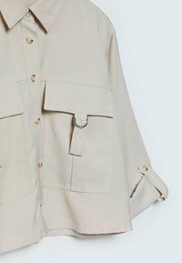 Chemise beige surdimensionnée avec col, deux poches avant, fermeture à boutons et manches retroussées. Confectionnée dans un tissu doux et lisse.